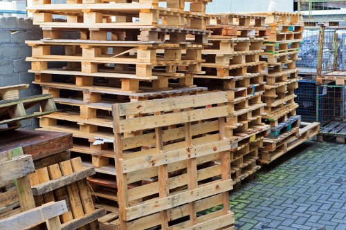 A large stack of wooden pallets and crates arranged in an outdoor storage area, situated on a paved surface with a brick wall on the left and a chain-link fence on the right. The pallets are made from light-colored, untreated timber, with some showing visible grain and rough edges, stacked in an orderly manner to form a tall, dense pile. Smaller pallets and crates are positioned in front of the main stack, some leaning against each other. The background includes industrial fencing and part of a building structure, indicating a commercial or warehouse environment typical of rubbish removal sites in Dulwich. Natural sunlight illuminates the scene, highlighting the textures of the wooden surfaces and casting soft shadows on the ground. Business Waste Removal Dulwich often handles such wooden waste, which can be commonly seen in urban rubbish clearance operations around the area.
